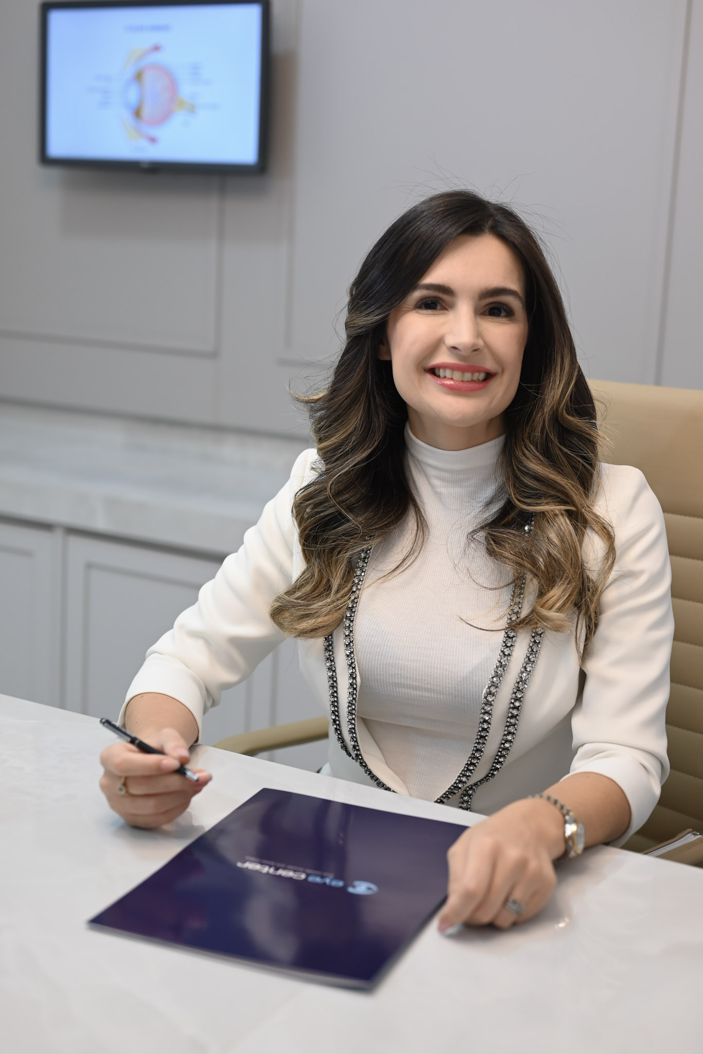 Retrato profissional da Dra. Ana Cristina Santiago Ribeiro, médica oftalmologista, jaleco branco, sorriso confiante e acolhedor, sentada em consultório elegante com mármore claro, iluminação suave, fotografia realista