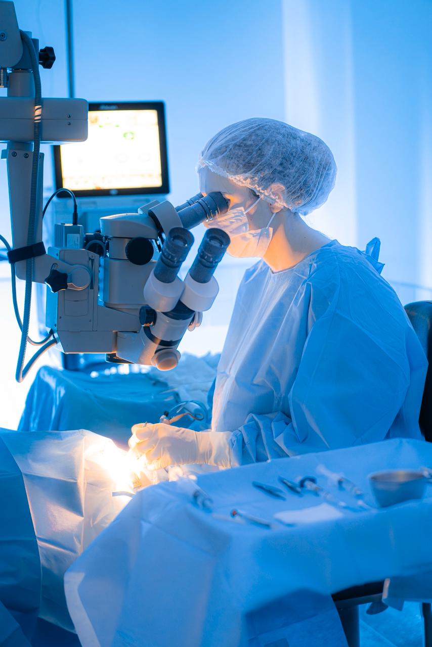 Cirurgião oftalmologista realizando procedimento cirúrgico de precisão utilizando microscópio cirúrgico em centro cirúrgico, ambiente asséptico em tons de azul, iluminação técnica intensa, paramentação completa, fotografia realista